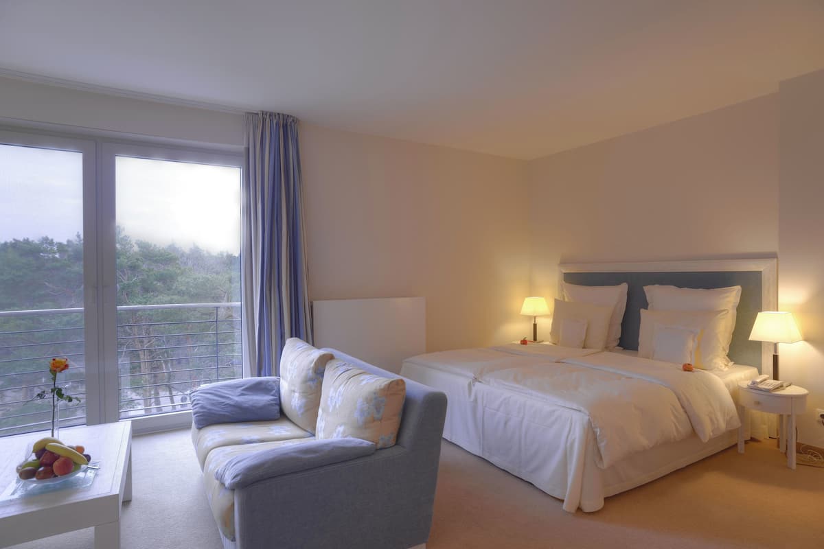 Ein Hotelzimmer mit Bett, einer Couch, einem Nachttisch, einer Lampe, einem Fenster mit Vorhängen und Blick auf Bäume.