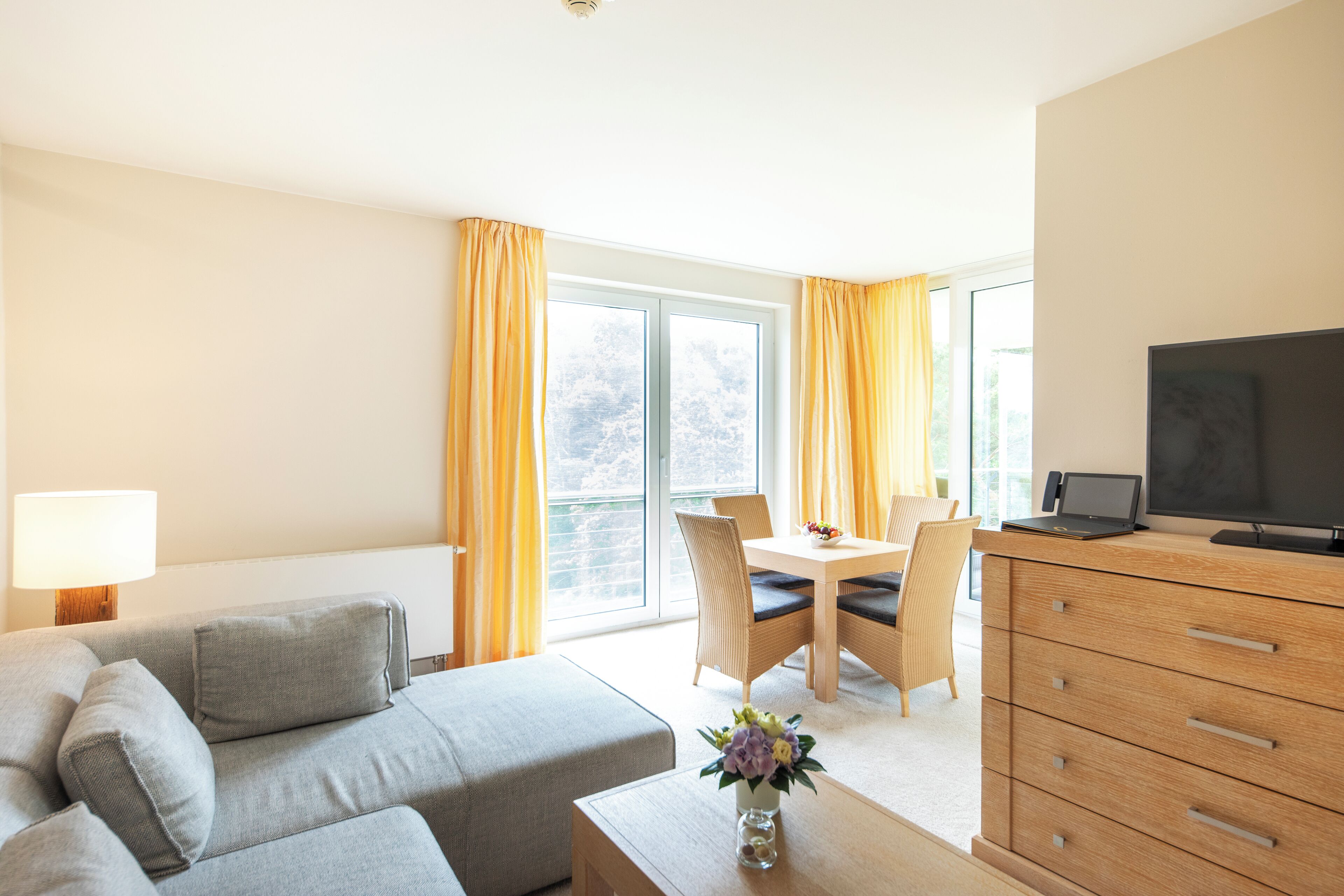 A modern hotel room with a sofa, a dining table with chairs, a TV, and a wooden dresser.