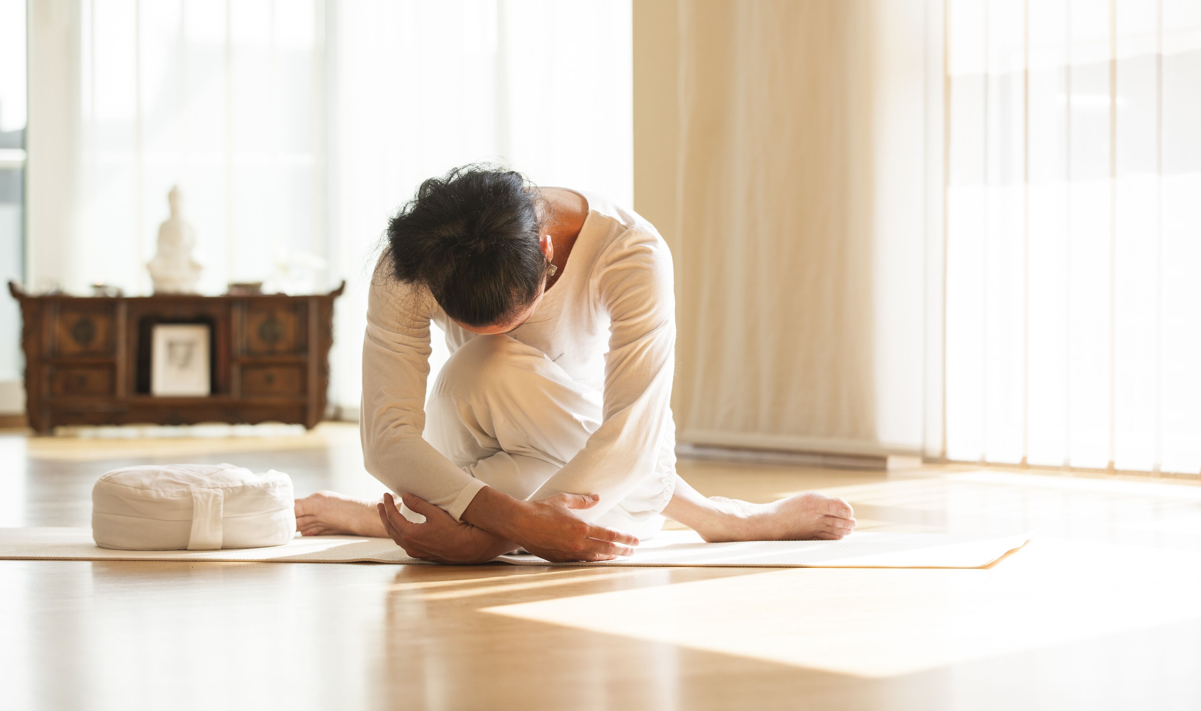 A person performing a yoga pose on a mat in a room with wooden flooring and a cabinet in the background.