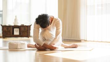 A person performing a yoga pose on a mat in a room with wooden flooring and a cabinet in the background.