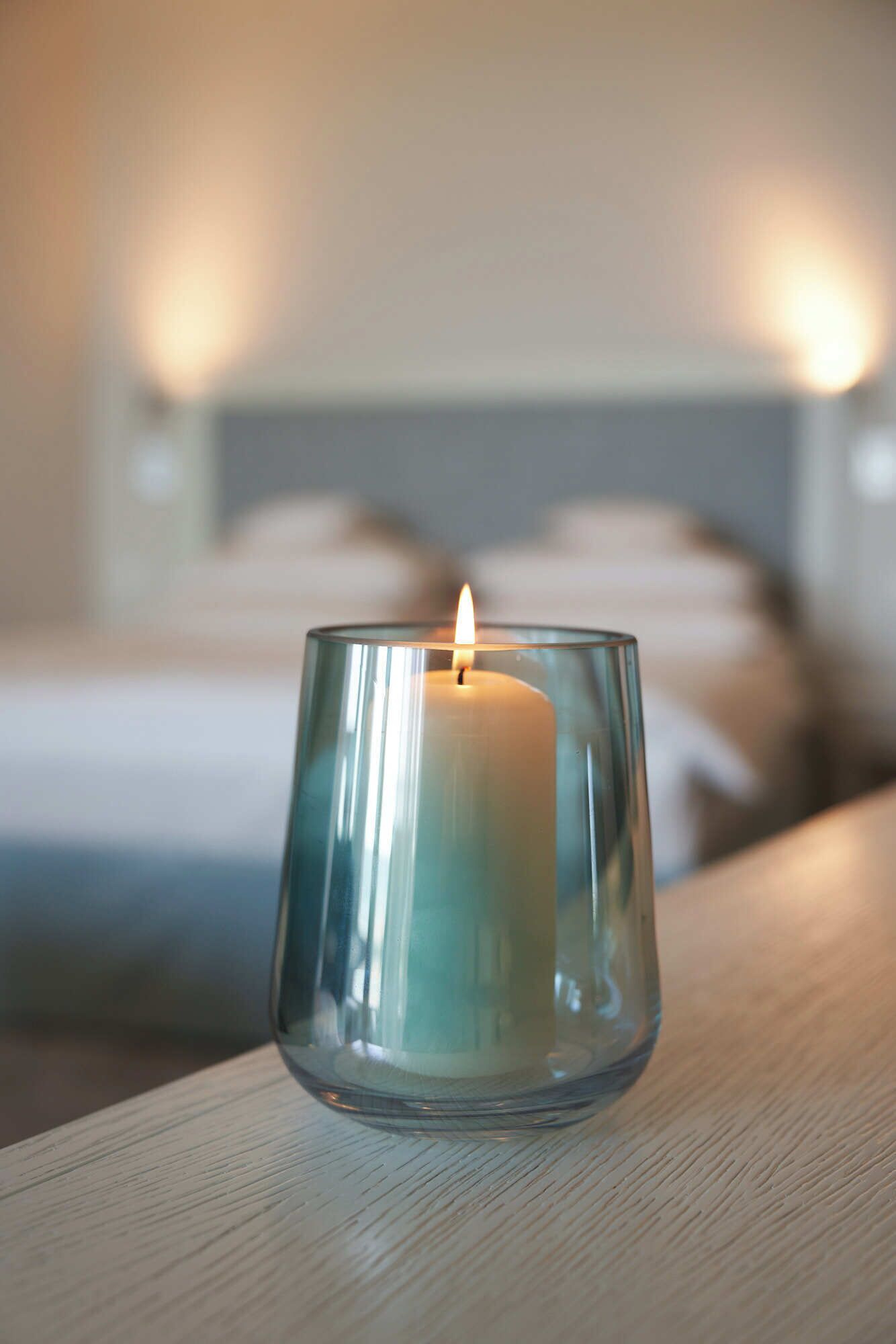 Una vela en un portaluz sobre una superficie de madera, con un fondo desenfocado de una cama y almohadas.