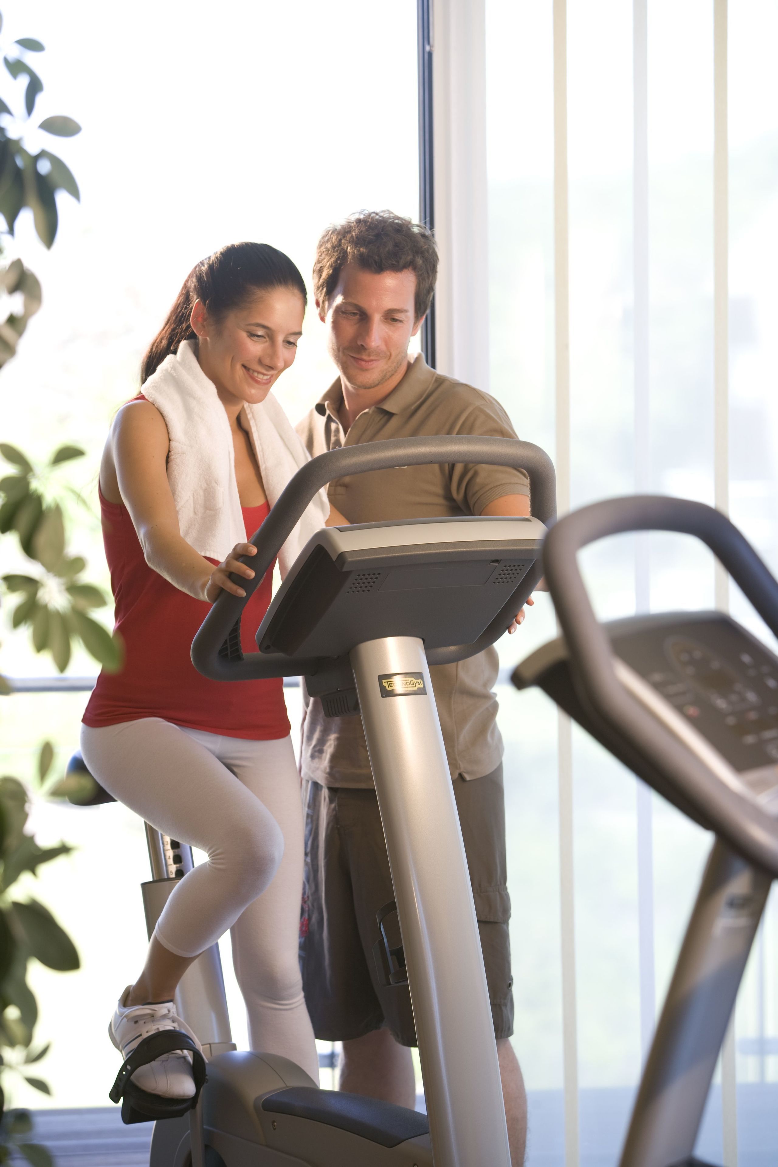A man and a woman using a treadmill in a gym with large windows and indoor plants.