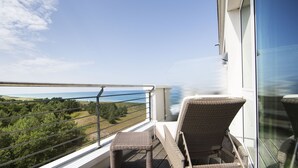 Un balcon doté de fauteuils en osier et d’une balustrade en verre, offrant une vue sur un paysage côtier.