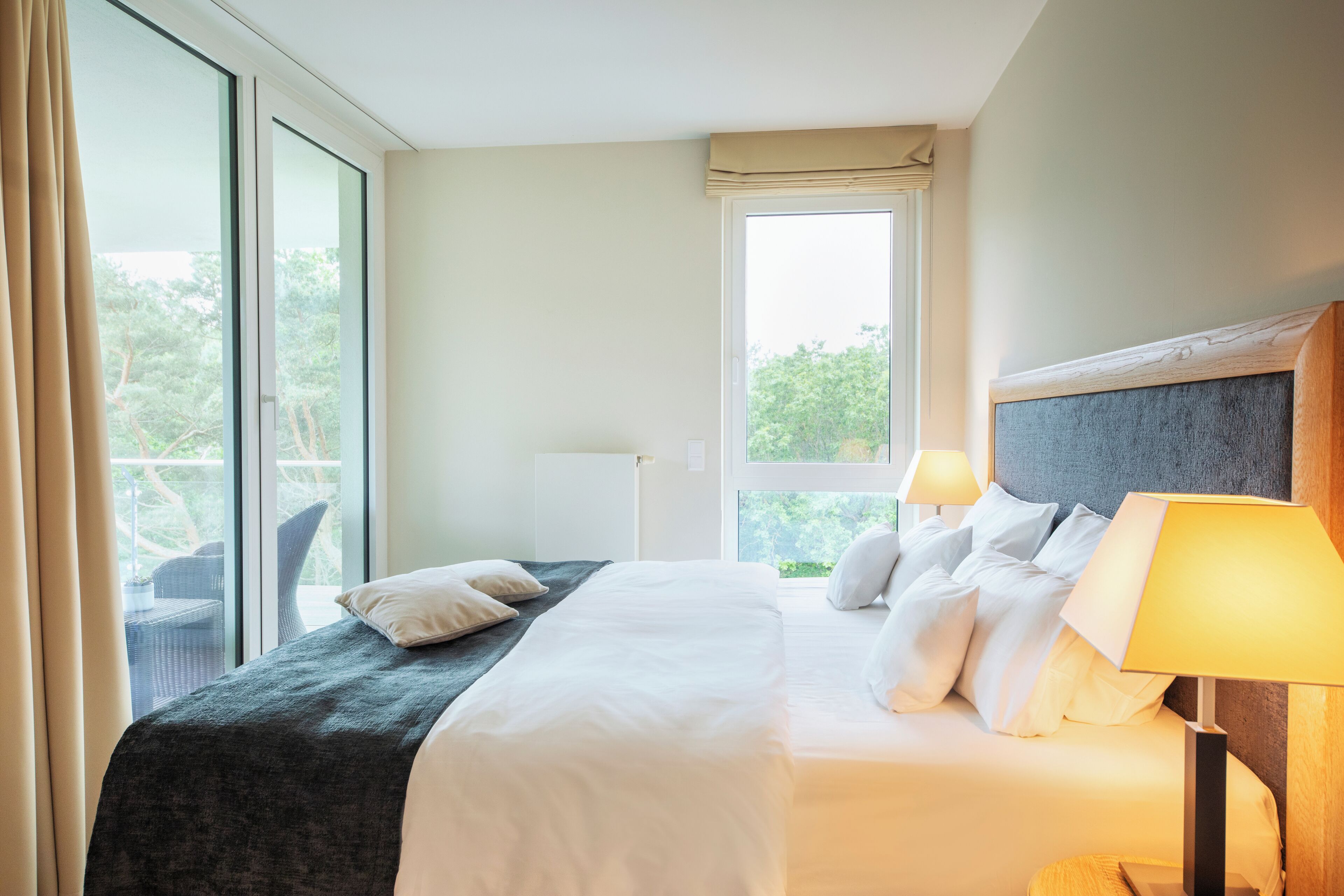 A hotel room with a large bed, a nightstand, a window with a view of trees, and a balcony with wicker furniture.