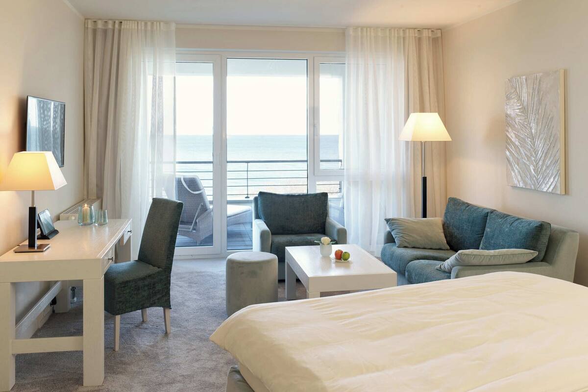 Ein Hotelzimmer mit einem Bett, zwei Sesseln, einer Couch, einem kleinen Tisch, einem Schreibtisch mit Lampe und Meerblick durch große Fenster.