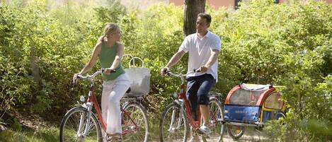 Dos personas montando en bicicleta por un sendero con vegetación y un edificio al fondo.