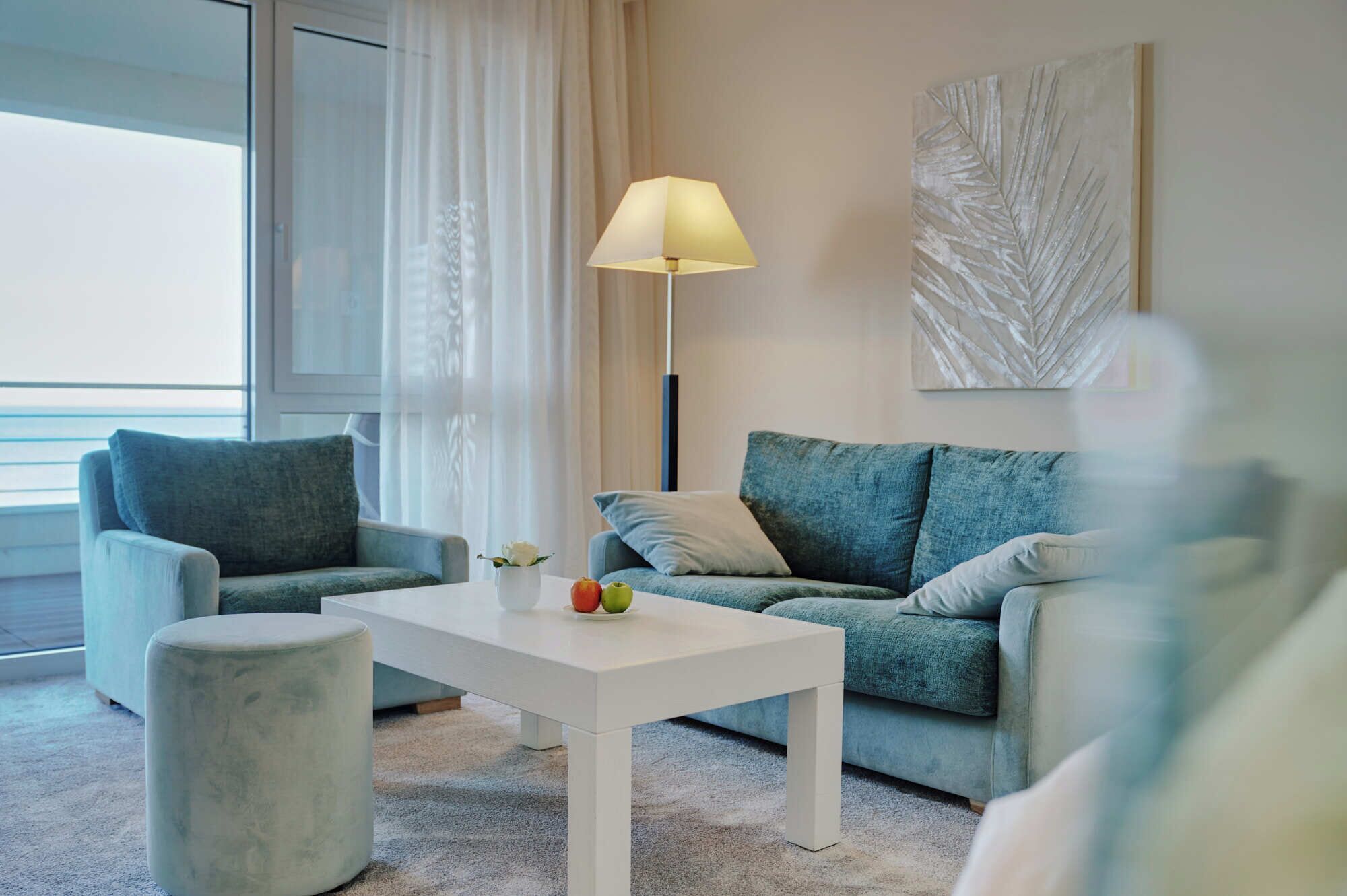 Ein modernes Wohnzimmer mit einem weißen Couchtisch, zwei grauen Sofas, einer Stehlampe und einem großen Fenster mit durchsichtigen Vorhängen.