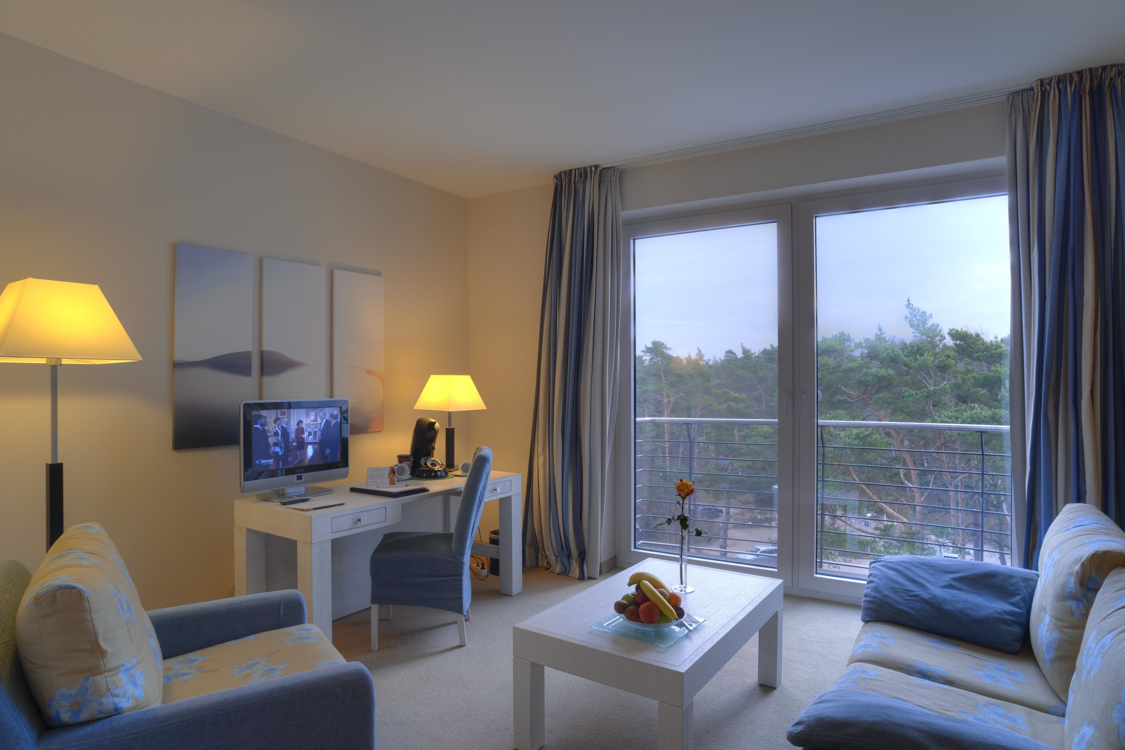 Habitación de hotel con escritorio, televisión, sofá, mesa de centro con fruta y balcón con cortinas.