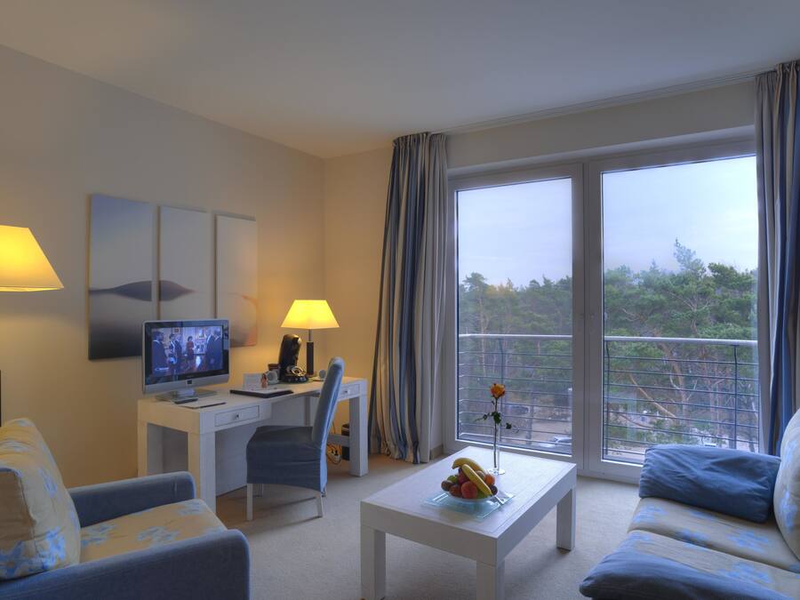 Habitación de hotel con escritorio, televisión, sofá, mesa de centro con fruta y balcón con cortinas.