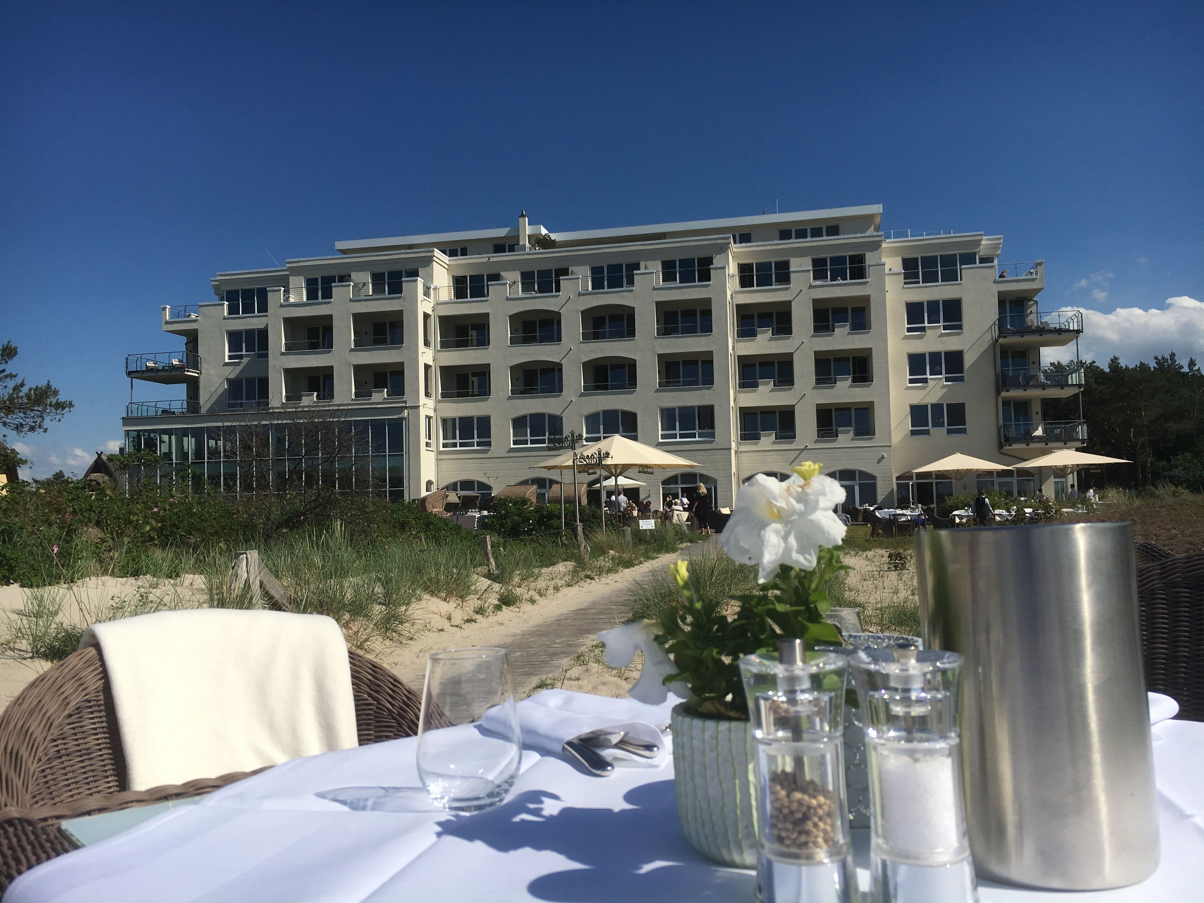 Een eettafel gedekt met een glas, zoutstrooiers en een metalen houder, met op de achtergrond een meerhoog gebouw en een strand.