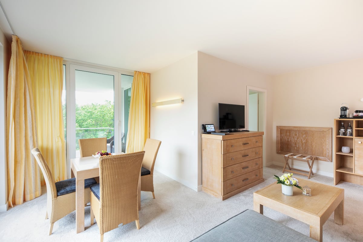 Ein modernes Hotelzimmer mit Balkon, einer Essecke mit Stühlen, einer Holzkommode, einem Fernseher und einem kleinen Tisch mit Blumen.