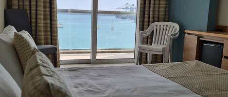 A hotel room with a bed, a desk, a chair, and a view of the sea and a bridge.