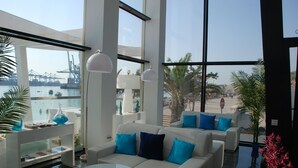 Ein modernes Wohnzimmer mit großem Fenster mit Blick auf einen Hafen, ausgestattet mit einem weißen Sofagarnitur, einem Glastisch und einer Stehlampe.