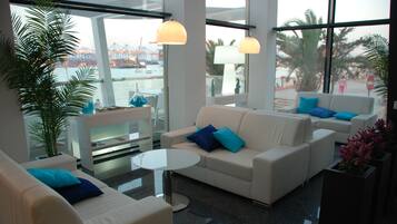 Un salon moderne avec des canapés blancs, une table basse en verre et de grandes fenêtres donnant sur un port.