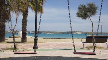 Un espacio con columpios frente a la playa, bancos, palmeras y vistas al mar.
