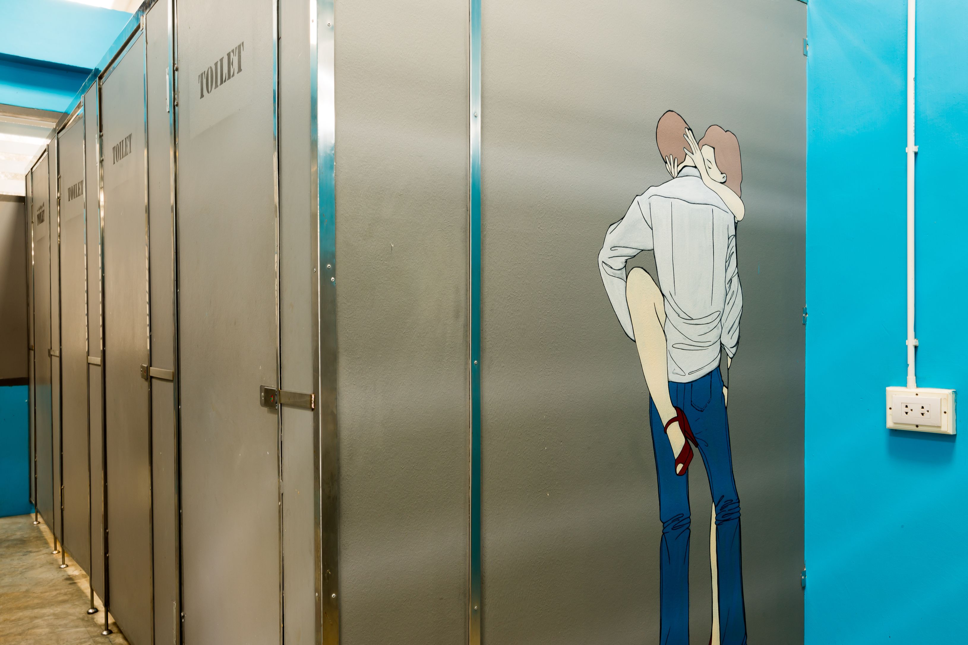 Una fila de cabinas de baño metálicas con una pared azul y una persona de pie frente a una de ellas.