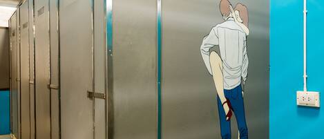 Una fila de cabinas de baño metálicas con una pared azul y una persona de pie frente a una de ellas.