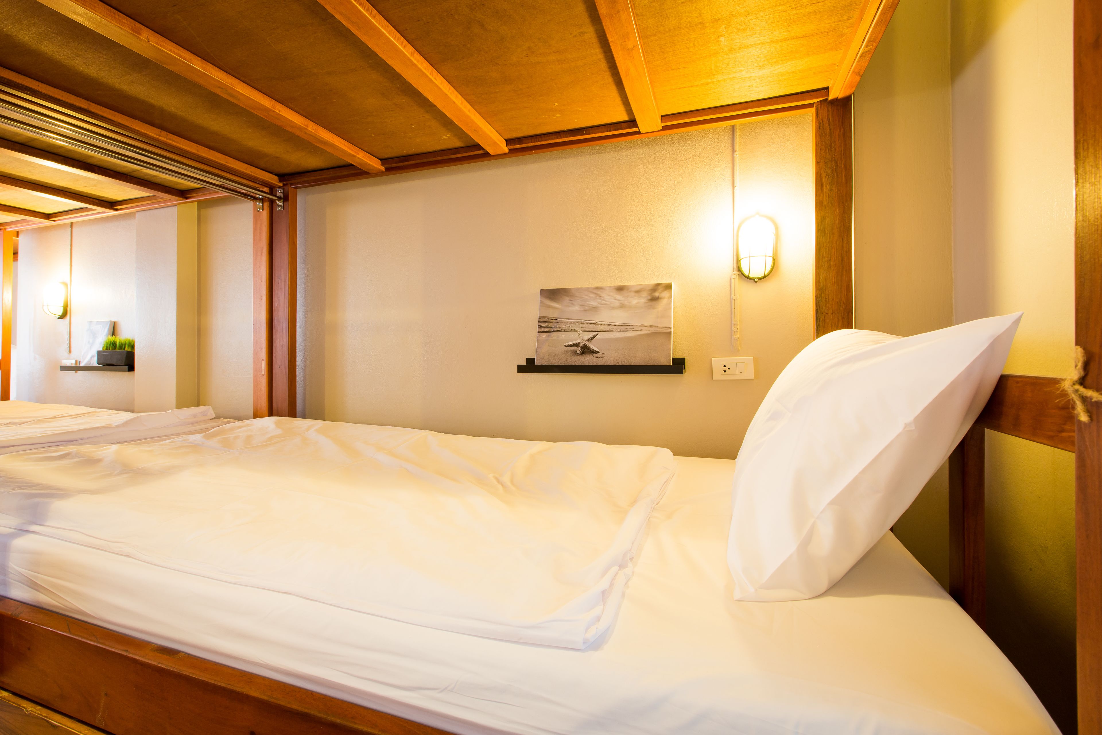Una cama bien hecha con sábanas blancas en una habitación con vigas de madera en el techo y una lámpara fijada a la pared.