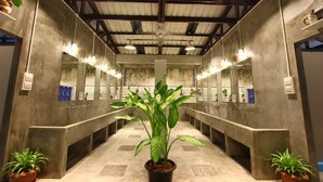 Een lange, smalle badkamer met meerdere wastafels en spiegels, potplanten en een hoog plafond met zichtbare balken.