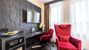 A modern hotel room with a dark marble wall, a flat-screen TV, a desk with a bowl of fruit, a red chair, and a large window with curtains. - PLAZA INN Salzburg City (Salzburg)