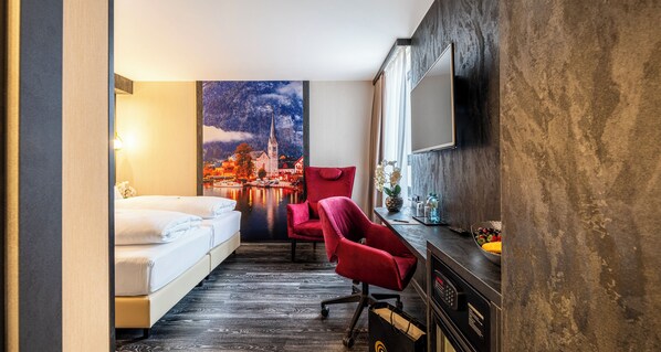 A hotel room with a bed, a red chair, a desk with a mirror, and a large wall mural of a cityscape.