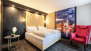 A modern hotel room with a large bed, a red chair, and a scenic mural of a town by a river. - PLAZA INN Salzburg City (Salzburg)