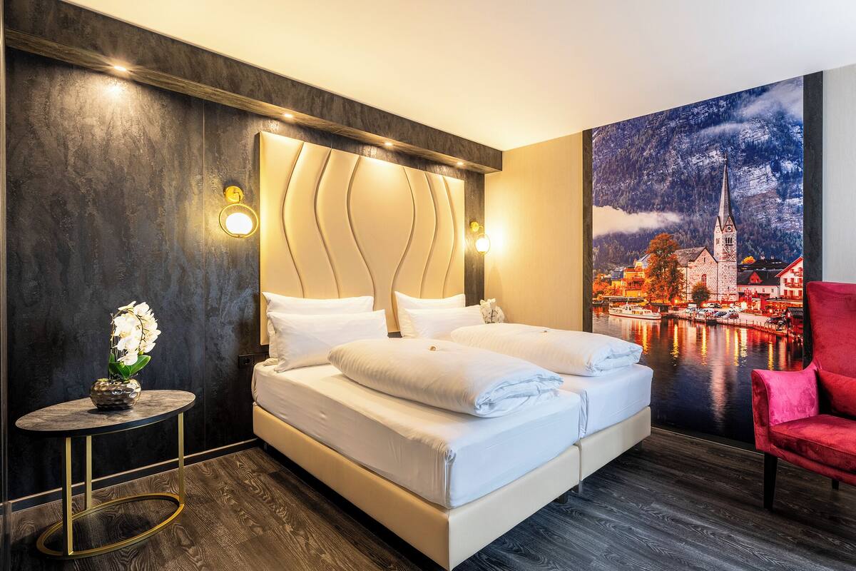 Une chambre d’hôtel moderne avec un grand lit, un fauteuil rouge et une fresque murale représentant un paysage urbain au bord d’une rivière.