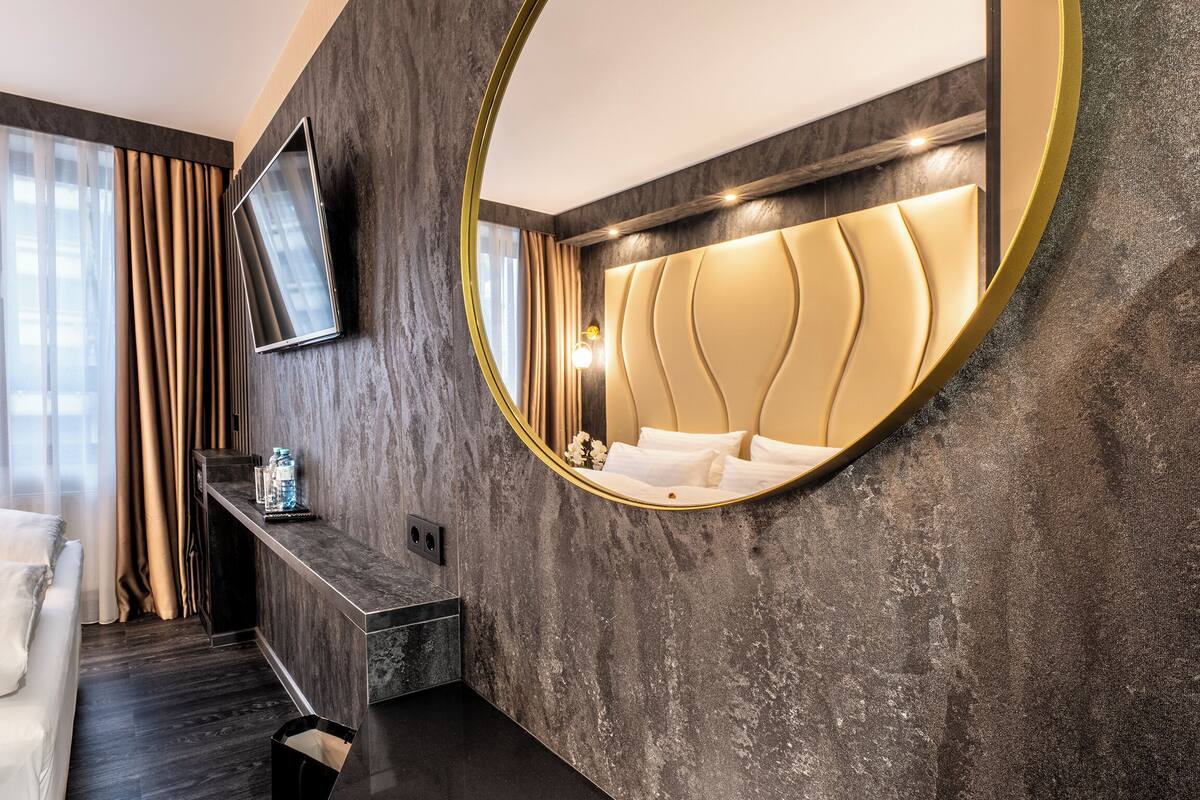Une chambre d’hôtel moderne équipée d’un lit, d’un téléviseur fixé au mur, d’un miroir rond et d’une banquette intégrée avec une petite table.
