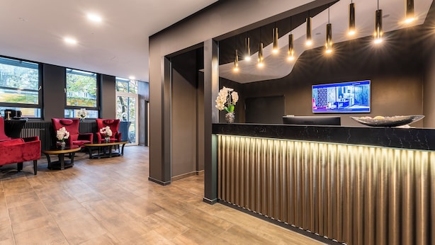 A modern reception area with a marble counter, a television, and a seating area with red chairs. - PLAZA INN Salzburg City (Salzburg)