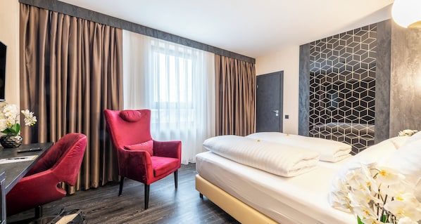 Habitación de hotel con cama, un sillón rojo, un escritorio y una silla, y un ventanal con cortinas.