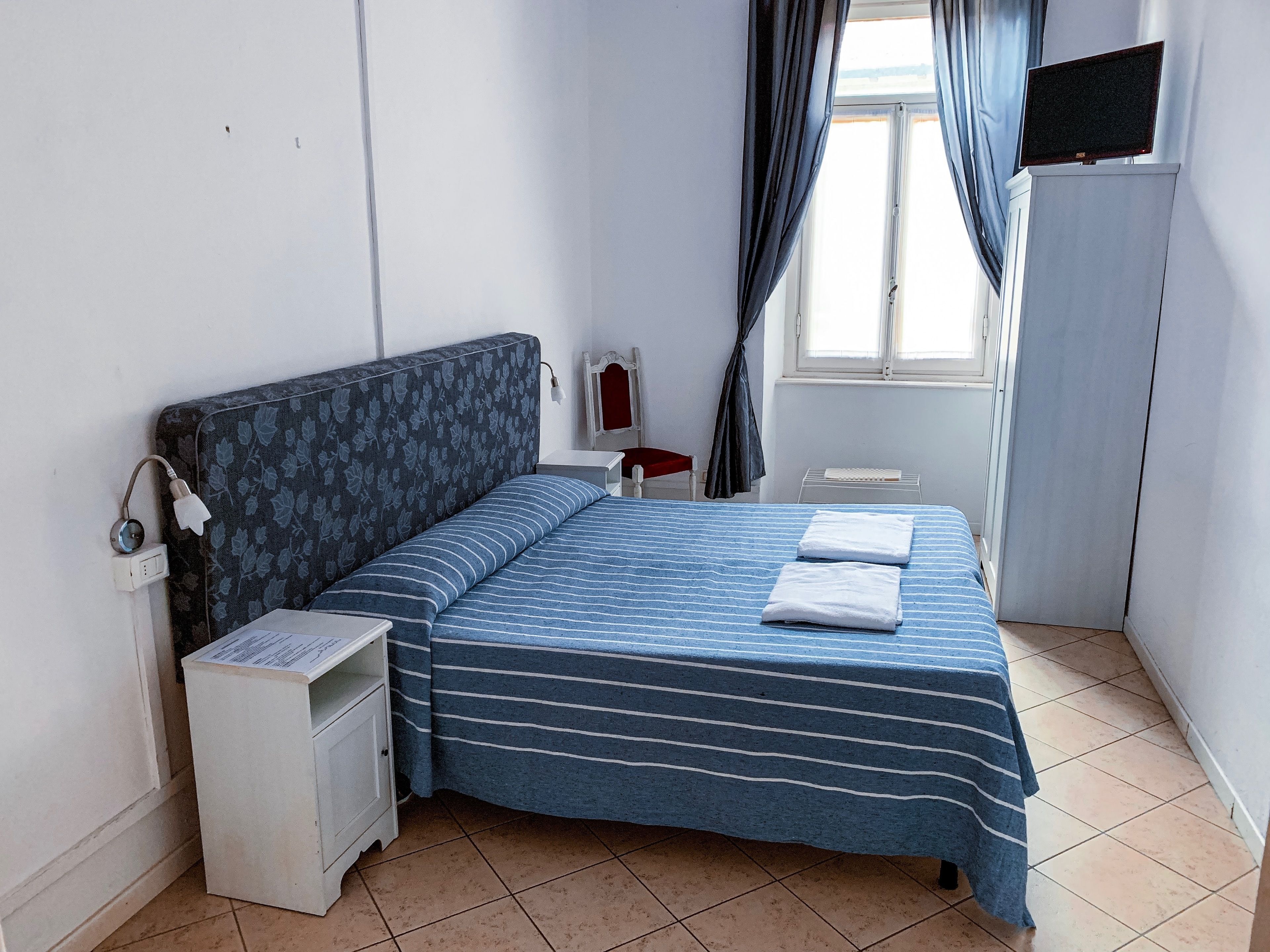 Une chambre à coucher comprenant un lit, une table de chevet, un bureau, une chaise, une fenêtre avec des rideaux et un téléviseur fixé au mur.