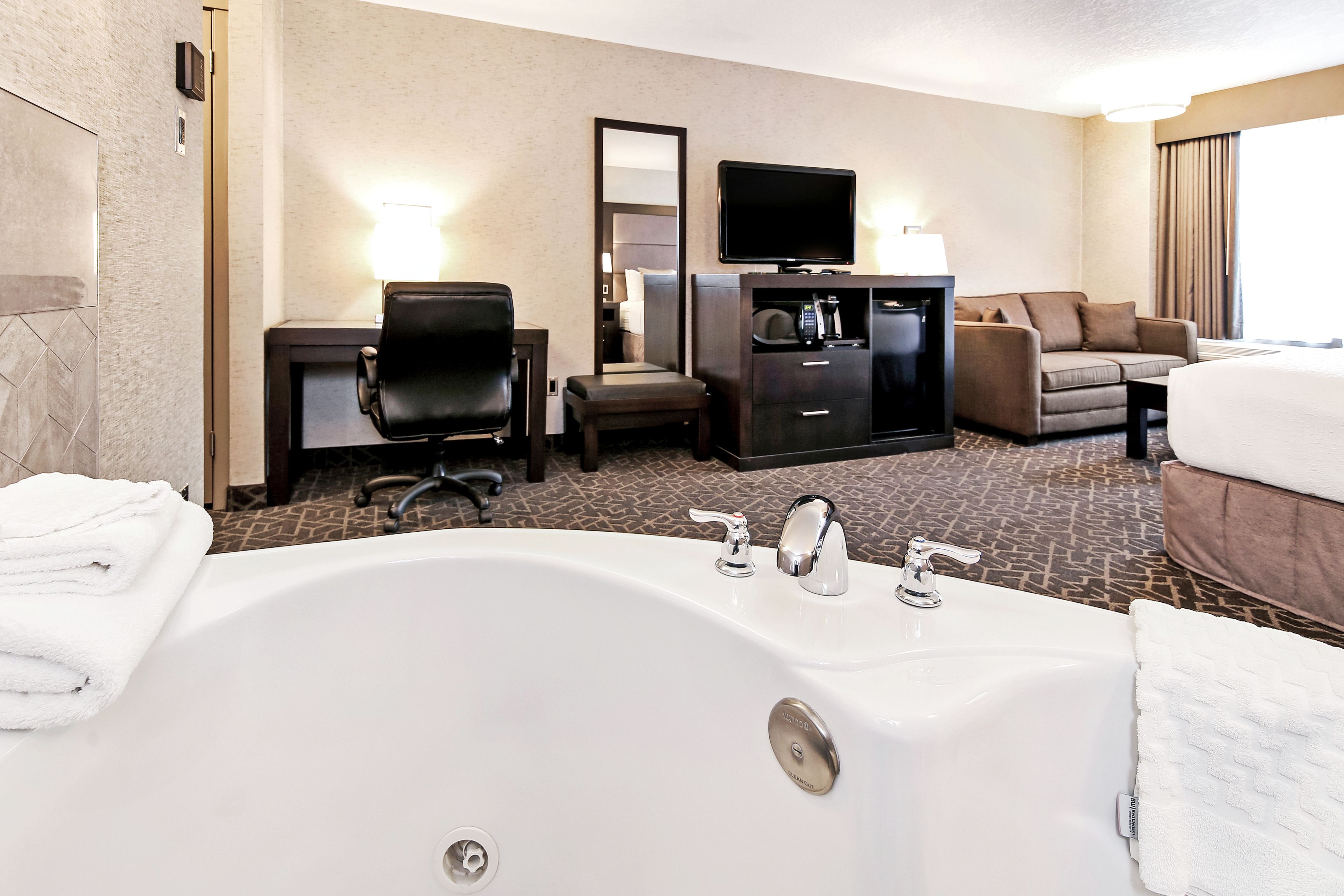 A hotel room with a large white bathtub, a sofa, a desk with a flat-screen TV, and a minibar.
