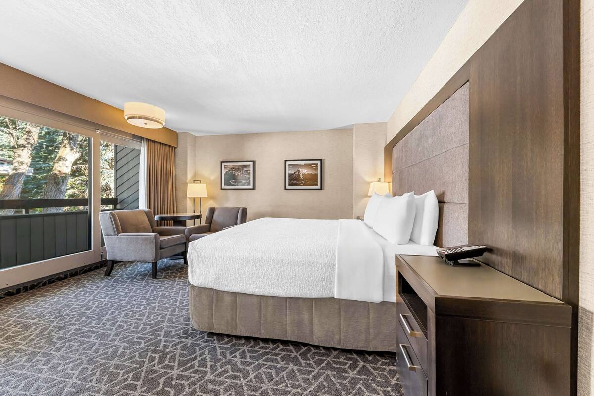 A hotel room with a large bed, a desk with a phone, two chairs, a window with curtains, and two framed pictures on the wall.