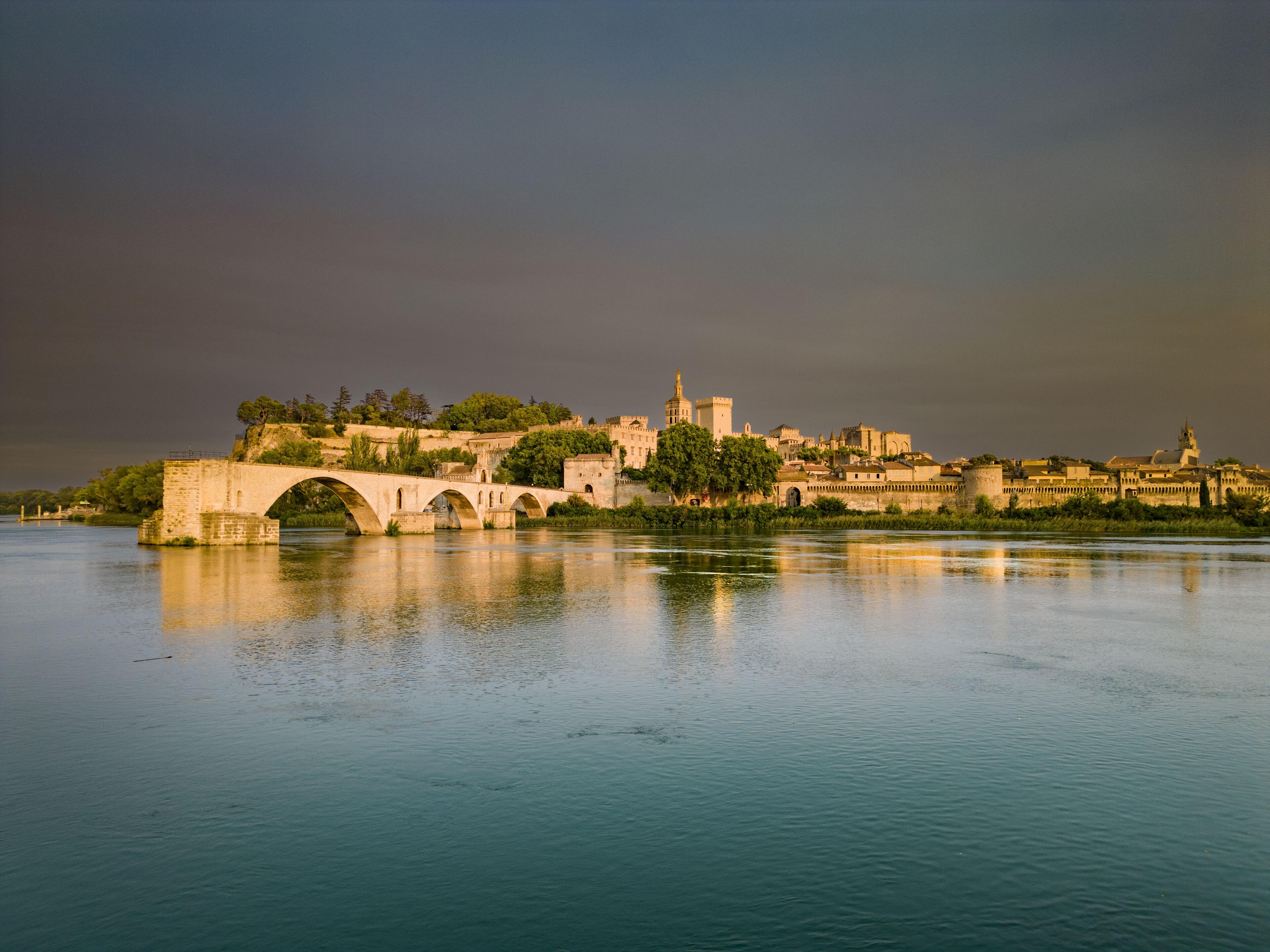 A historic bridge over a calm river, leading to a fortified city with ancient architecture.