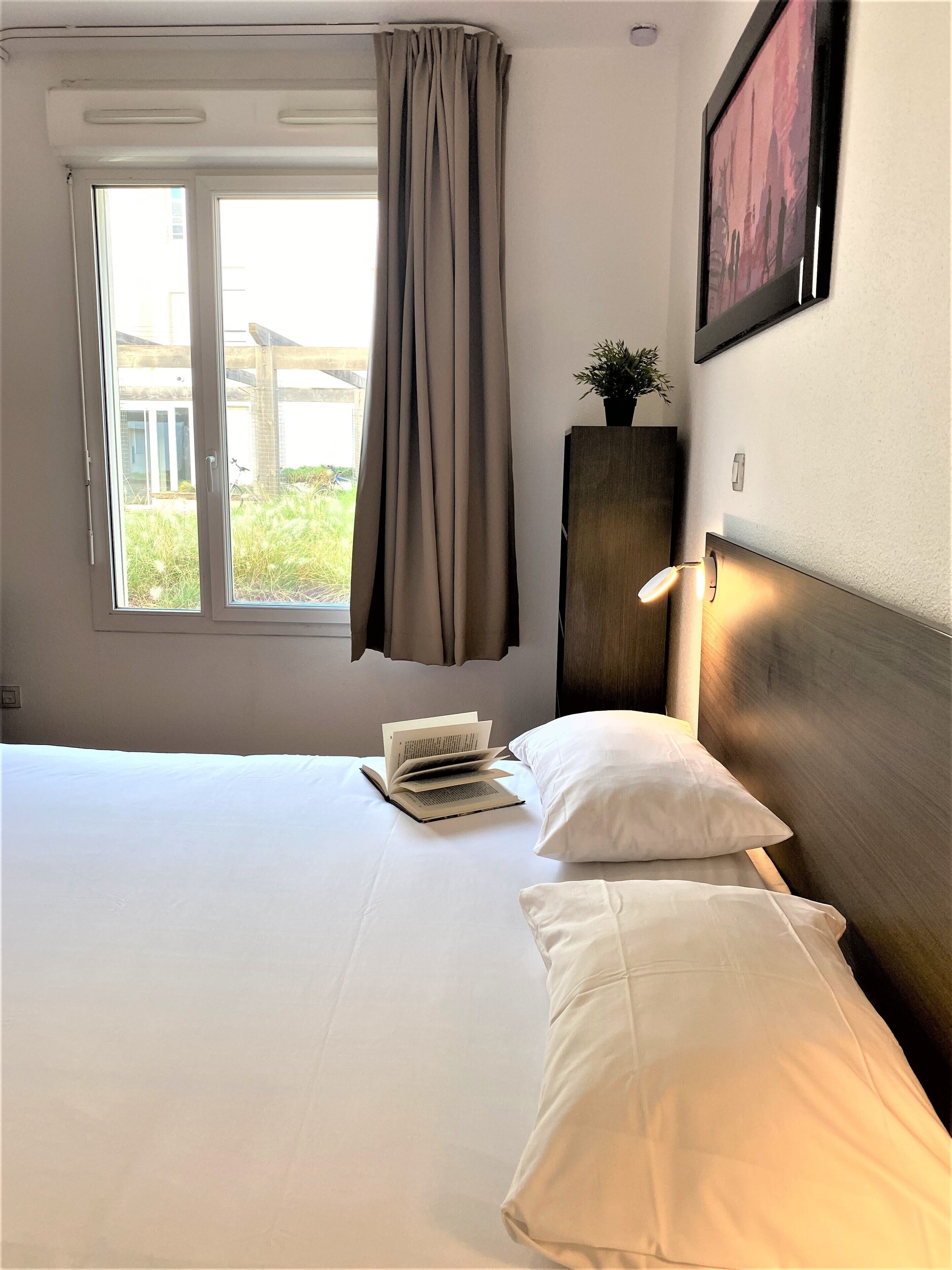Habitación de hotel con cama, ventana con cortinas, mesita de noche con un libro, planta en maceta y lámpara de pared.