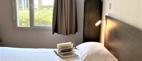 Une chambre d’hôtel avec un lit, une fenêtre avec des rideaux, une table de chevet avec un livre, une plante en pot et une lampe fixée au mur.