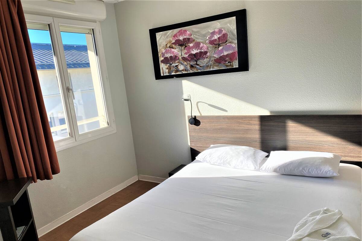 A hotel room with a bed, a window with curtains, a painting of flowers, and a wooden headboard.