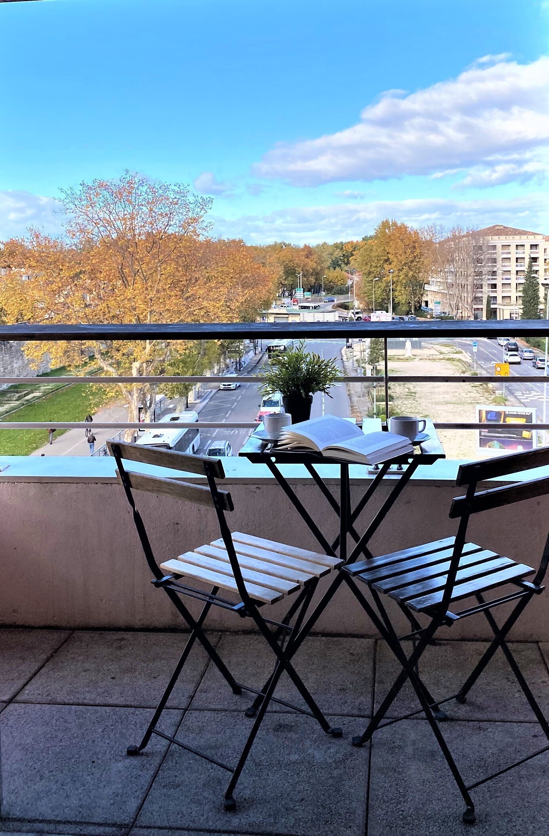 Un balcón con una mesa, sillas, una planta en maceta y vistas a una calle y edificios de la ciudad.