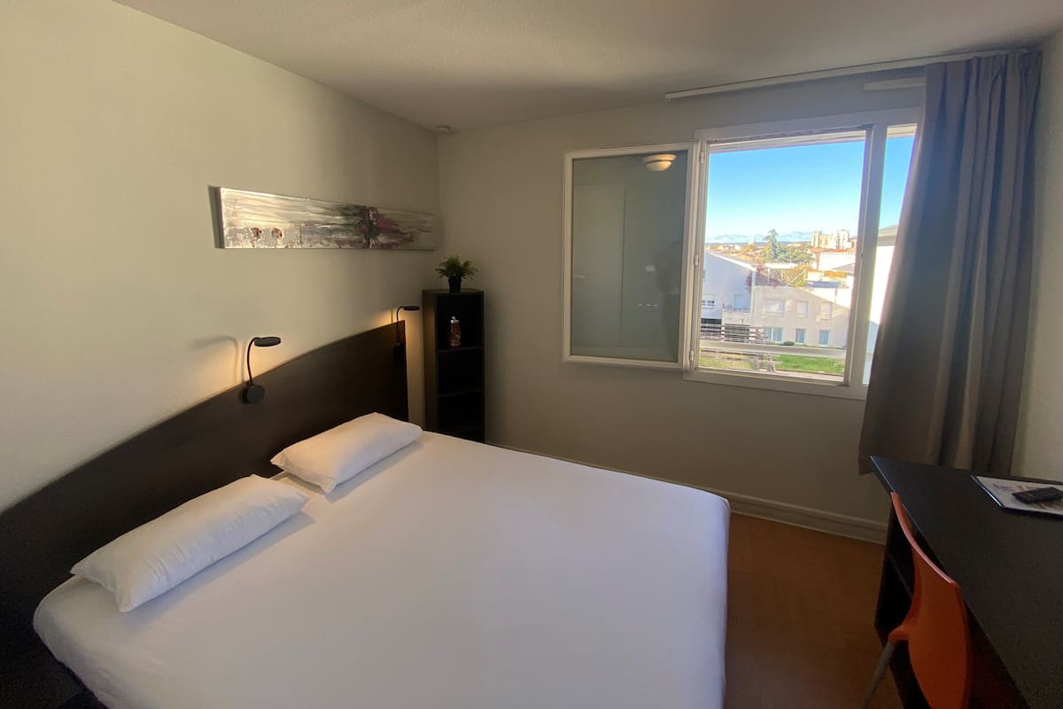 A hotel room with a bed, a desk, a window with a view of buildings, and a wall-mounted shelf with a plant.