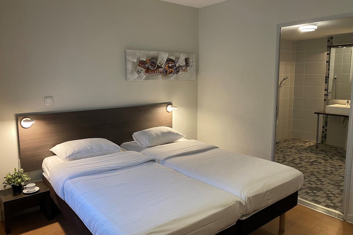 A double bed with white linens, a nightstand with a cup and a plant, a wall-mounted artwork, and a shower area with tiled walls.