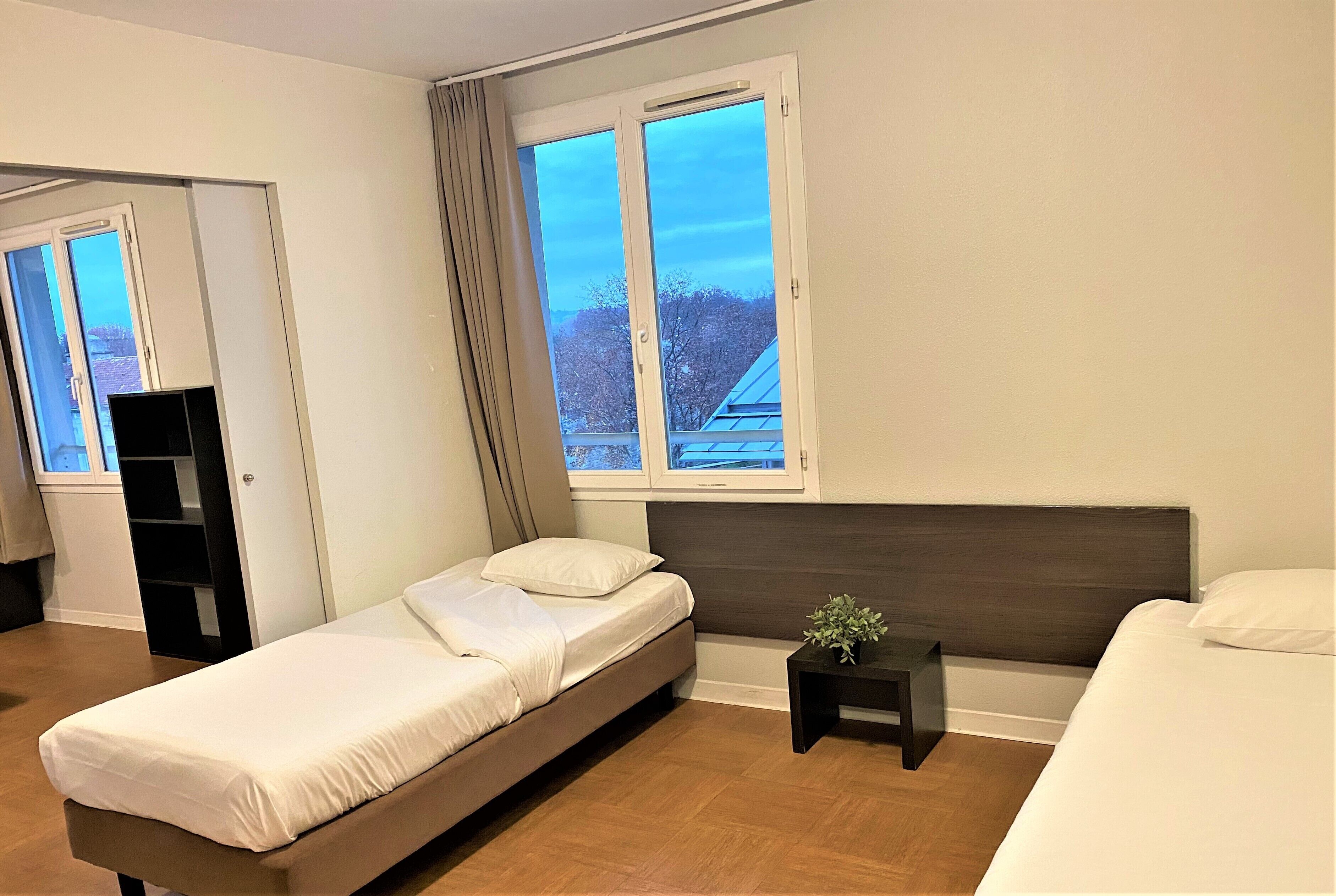 Une chambre à coucher moderne avec un lit simple, une table de chevet, une petite table avec une plante et une fenêtre avec des rideaux.