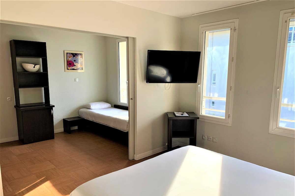 A bedroom with a bed, a TV mounted on the wall, a small bedside table, a window, and a shelf with a bowl.