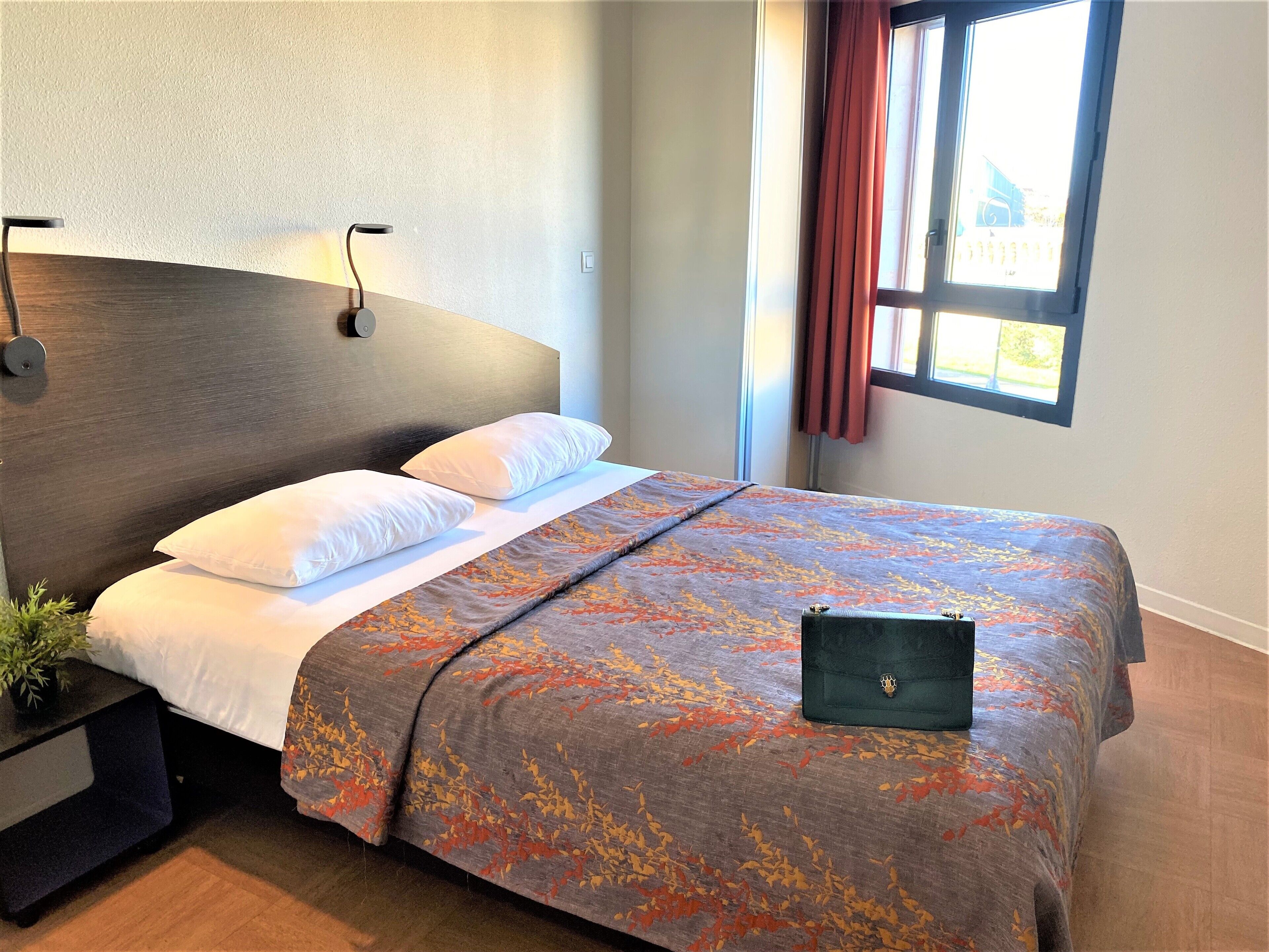 A neatly made bed with a patterned bedspread, a bedside table with a lamp, and a window with curtains.