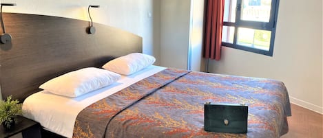 Un lit bien fait, recouvert d’une couvre-lit à motifs, une table de chevet avec une lampe et une fenêtre avec des rideaux.