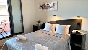 Een opgemaakt bed met twee opgerolde handdoeken, een nachtkastje met een lamp, een kleine bloempot en een kunstwerk aan de muur.