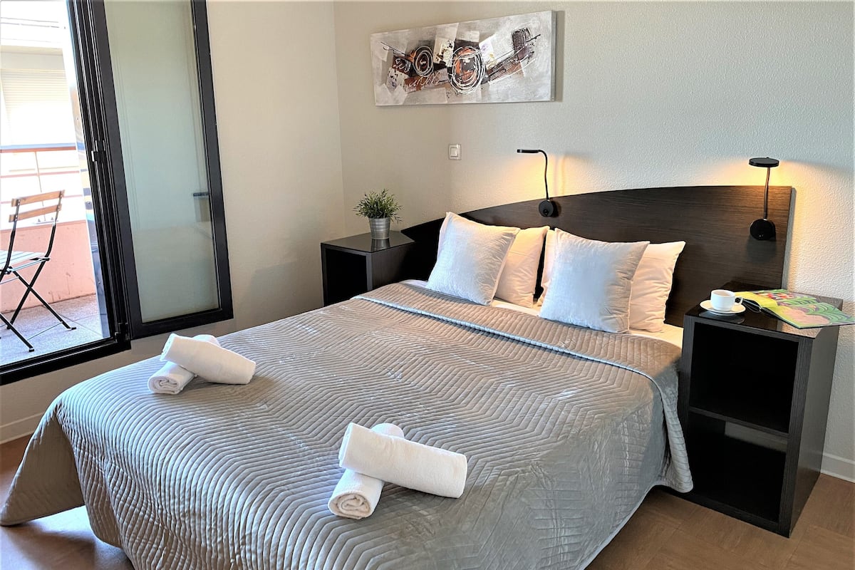 A neatly made bed with two rolled towels, a bedside table with a lamp, a small potted plant, and a wall-mounted artwork.