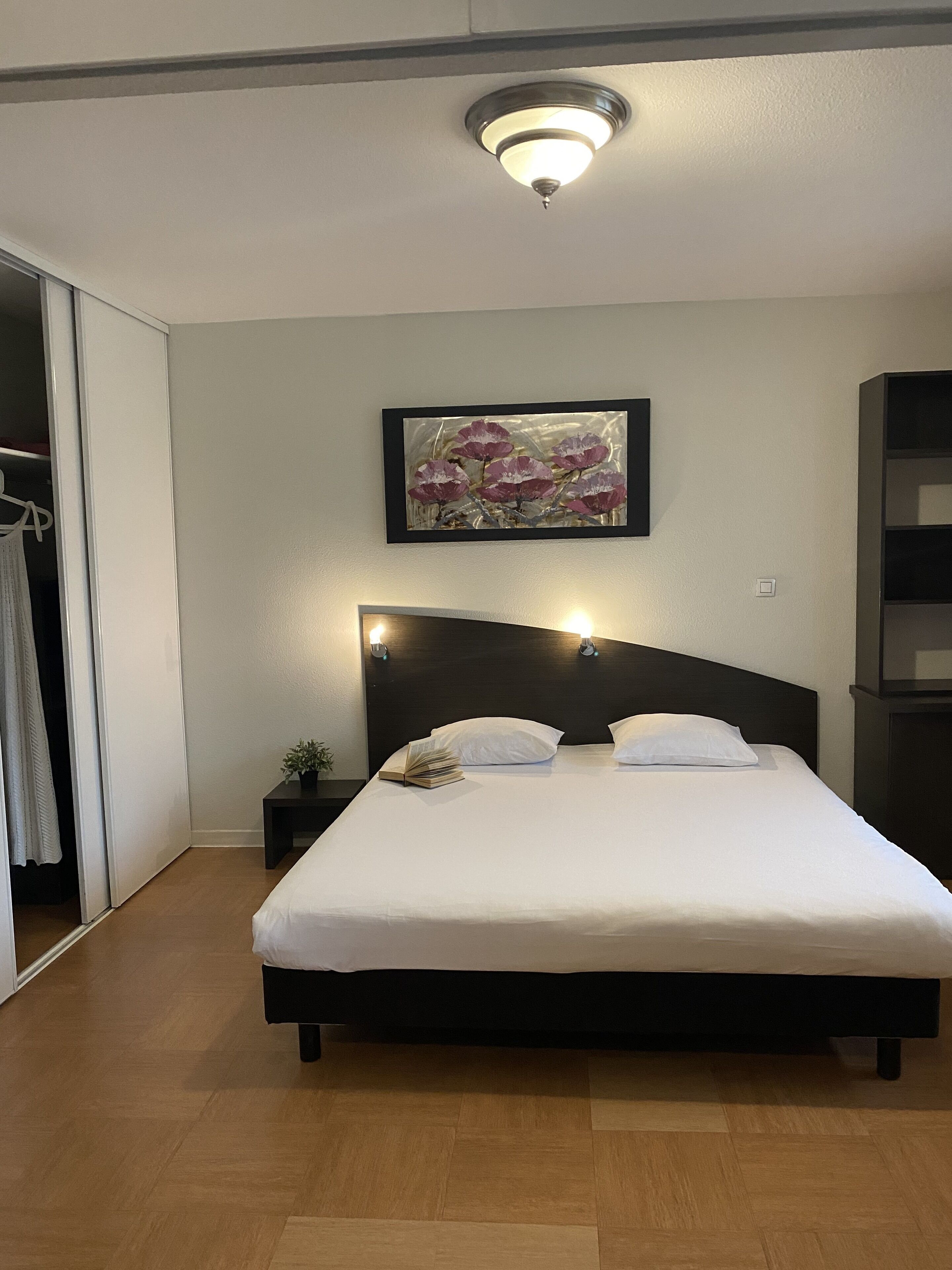 A neatly made bed with white linens, a dark headboard, and a bedside table with a plant.