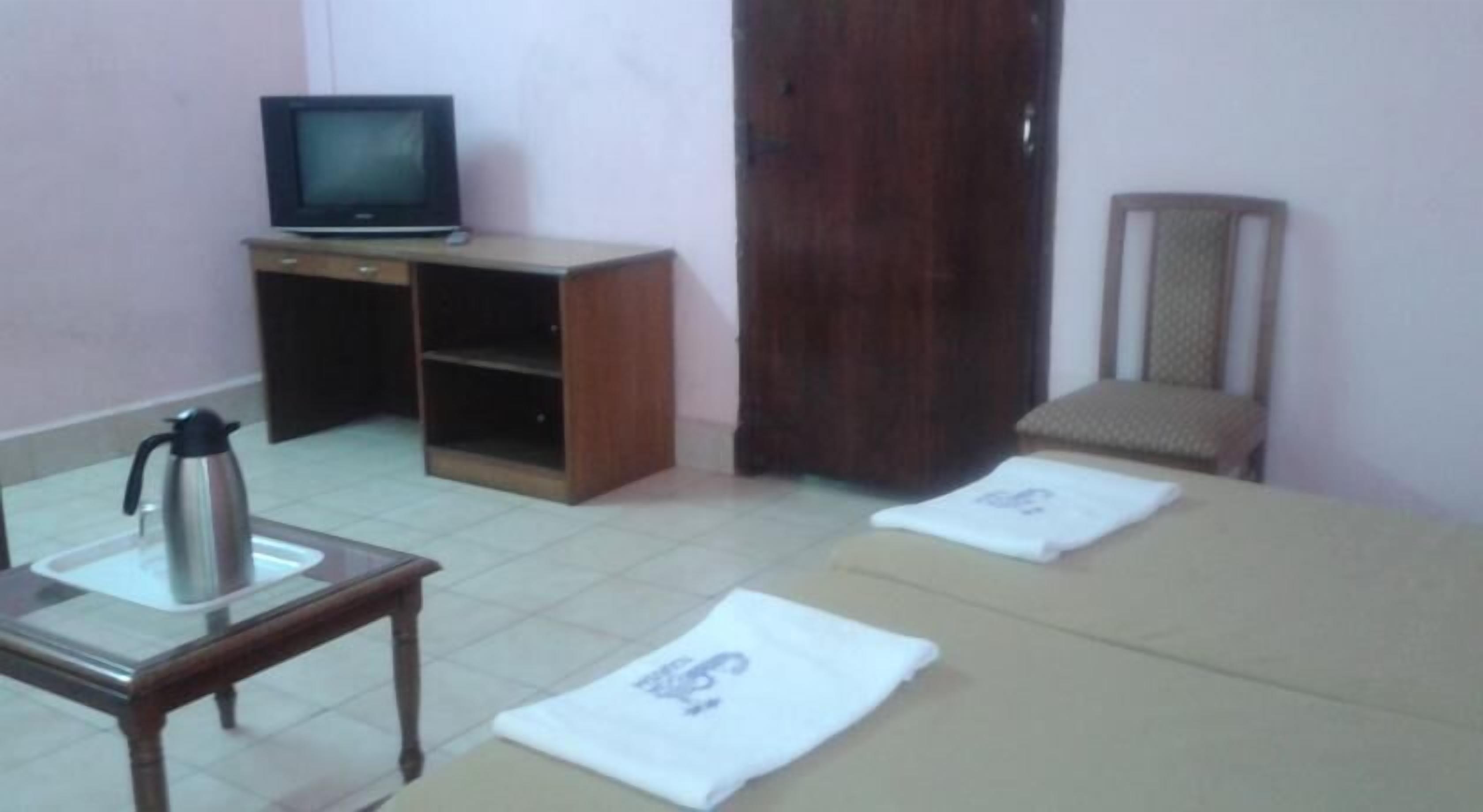 Quarto com cama, televisão sobre suporte de madeira, cafeteira e uma cadeira.