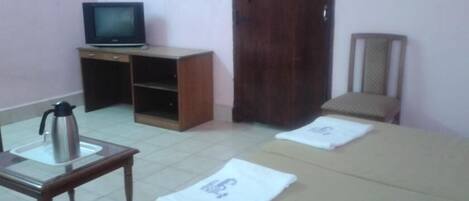 Quarto com cama, televisão sobre suporte de madeira, cafeteira e uma cadeira.