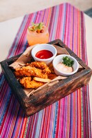Una bandeja de madera con pollo frito, papas fritas y dos salsas para mojar sobre un mantel colorido a rayas.